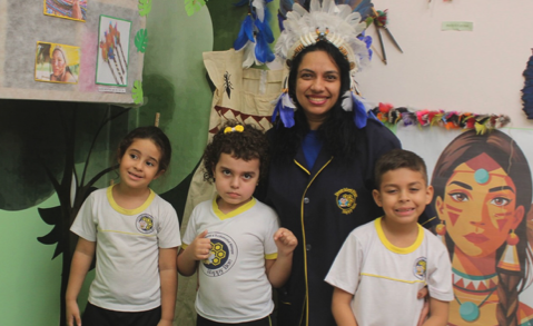Professora com cocar azul e três alunos em frente a painéis indígenas